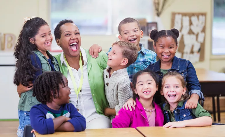 Une enseignante et sept jeunes élèves sourient et rient en posant ensemble dans une salle de classe.