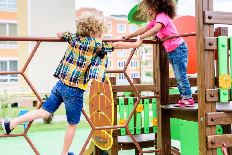 Deux enfants jouent sur une structure de jeux. L'un escalade un filet hexagonal, tandis que l'autre, debout sur une plateforme, tend la main. On aperçoit en arrière-plan des équipements de jeux colorés et des immeubles.