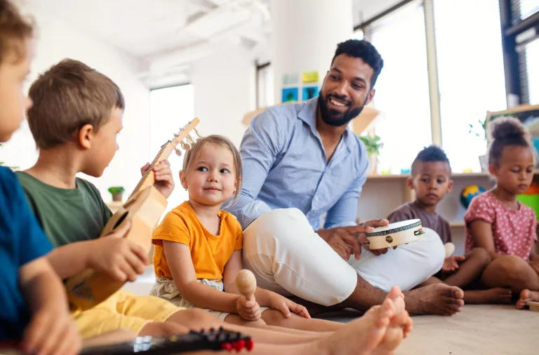Un groupe de jeunes enfants sont assis par terre avec des instruments de musique, dont un tambourin, une guitare et un maracas, tandis qu'un adulte est assis avec eux, souriant et interagissant avec eux.