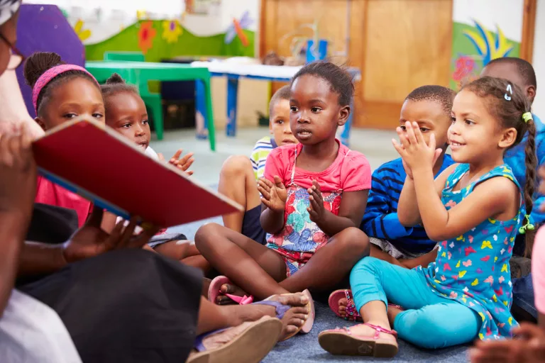 Dans une salle de classe colorée, un groupe d'enfants est assis par terre, applaudissant et écoutant un adulte lire un livre.