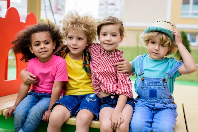 Quatre enfants vêtus de vêtements colorés sont assis côte à côte sur une structure de jeu à l'extérieur, souriant à l'objectif.